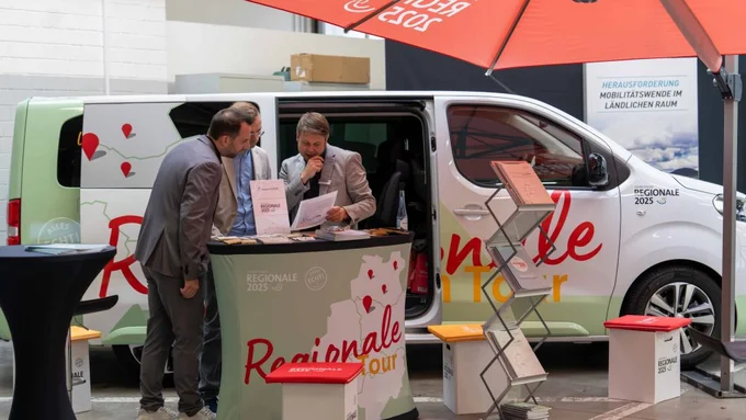 Drei Männer im Gespräch an einem Infostand mit REGIONALE-Branding vor einem Van mit rotem Sonnenschirm.