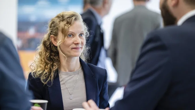 Frau mit lockigem Haar, dunkelblauem Blazer und hellbeigem Oberteil hält einen Pappbecher, im Hintergrund mehrere Personen in Gesprächssituation auf dem innocam.FORUM.