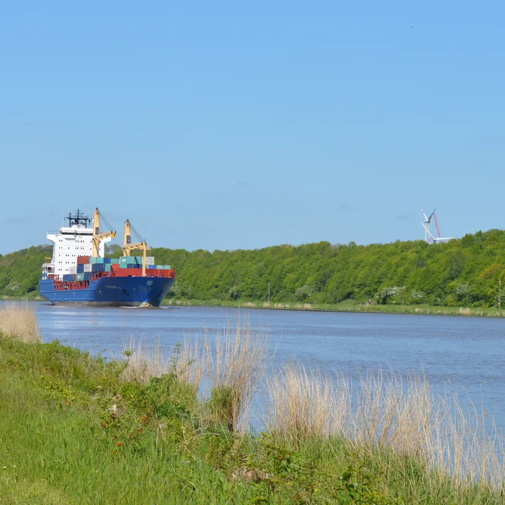 Frachtschiff fährt auf einem breiten Kanal neben einem Weg mit Gras und Bäumen unter klarem Himmel