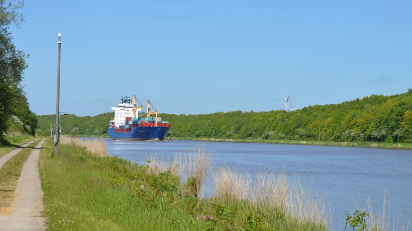 Frachtschiff fährt auf einem breiten Kanal neben einem Weg mit Gras und Bäumen unter klarem Himmel