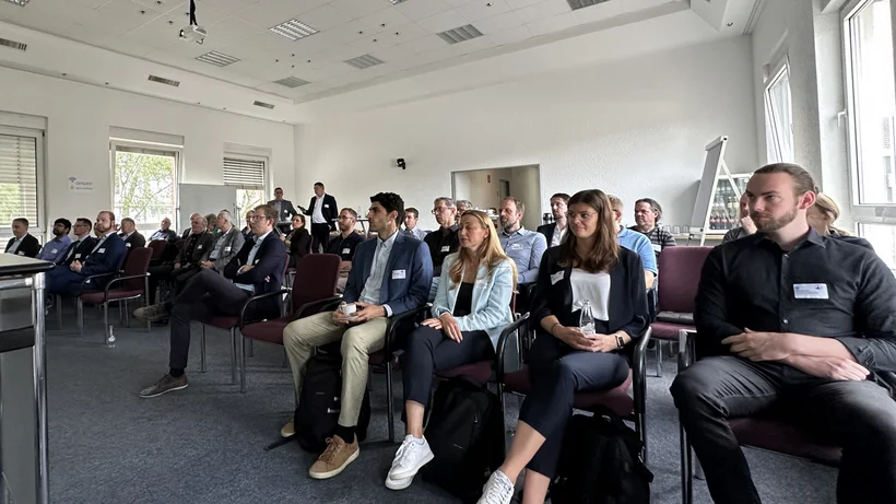 Eine Gruppe von Personen sitzt in einem Konferenzraum und hört einer Präsentation zu. Die Teilnehmer tragen Namensschilder und sind in Reihen auf Stühlen angeordnet. Der Raum ist hell mit großen Fenstern, durch die Tageslicht einfällt.