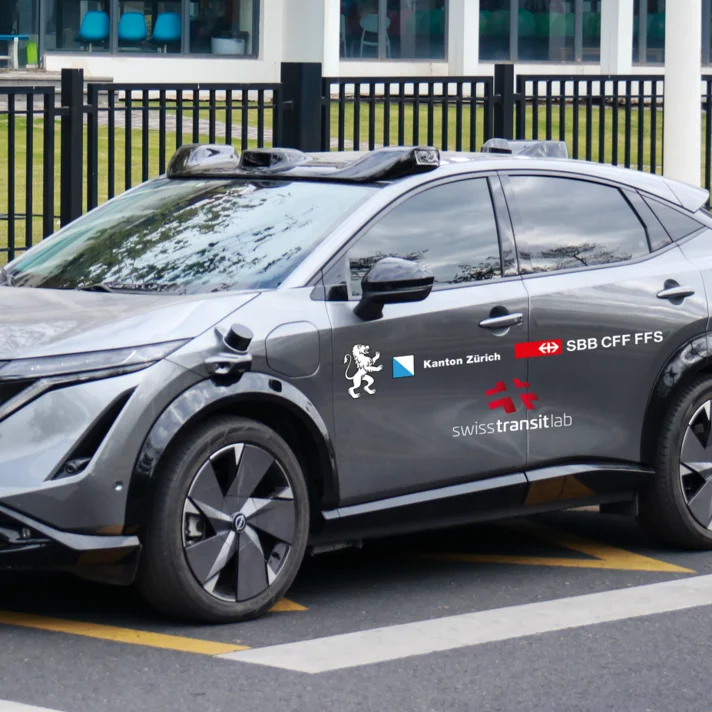 Ein modernes, graues Auto mit Sensoren auf dem Dach steht am Straßenrand. Logos mehrerer Projektpartner sind zu sehen.