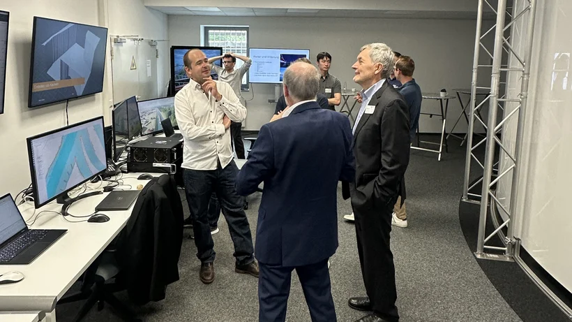 Acht Personen stehen in einem Raum mit vielen Monitpren und unterhalten sich.