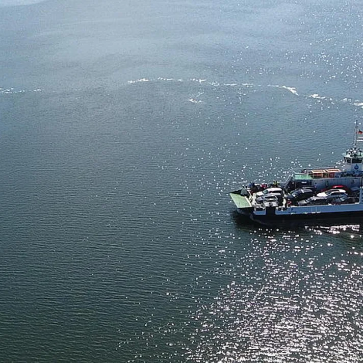 Luftaufnahme einer Autofähre mit mehreren Fahrzeugen an Bord auf offenem Gewässer, kleineres Boot im Hintergrund, glitzernde Wasseroberfläche.