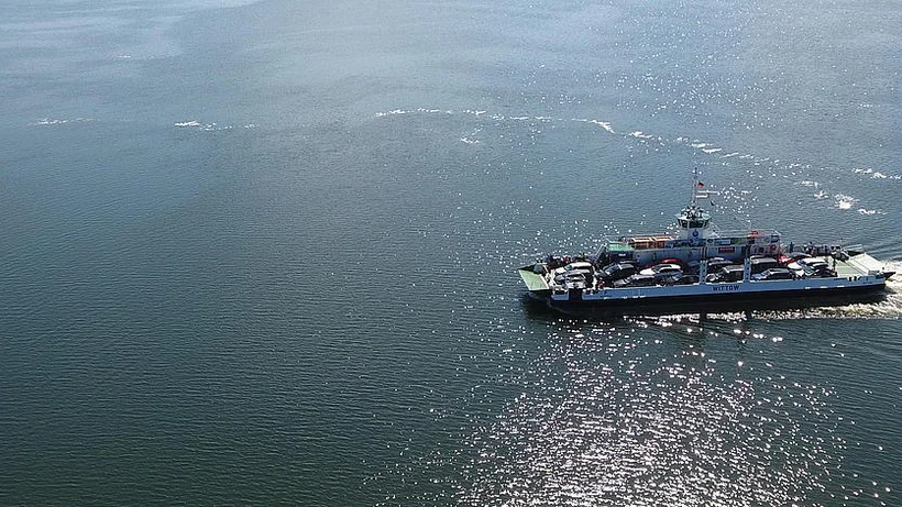 Luftaufnahme einer Autofähre mit mehreren Fahrzeugen an Bord auf offenem Gewässer, kleineres Boot im Hintergrund, glitzernde Wasseroberfläche.