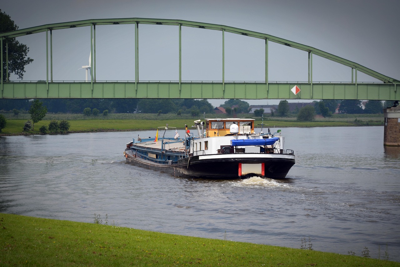 Binnenschiff mit weißem Aufbau und blauem Sonnenschutz fährt unter einer grünen Stahlbrücke auf einem Fluss.