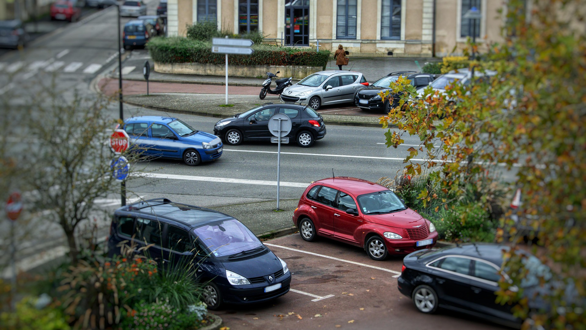 Blick auf eine belebte Straßenkreuzung mit mehreren parkenden und fahrenden Autos sowie einem Fußgänger auf dem Bürgersteig.