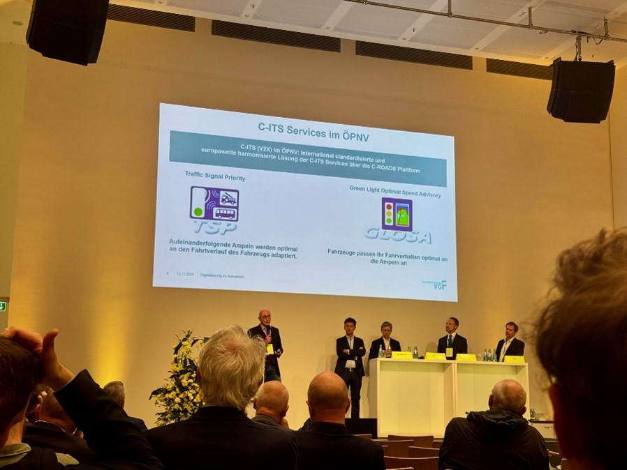 Podiumsdiskussion mit fünf Personen auf einer Bühne vor einer Präsentationsfolie zum Thema C-ITS Services im öffentlichen Nahverkehr.