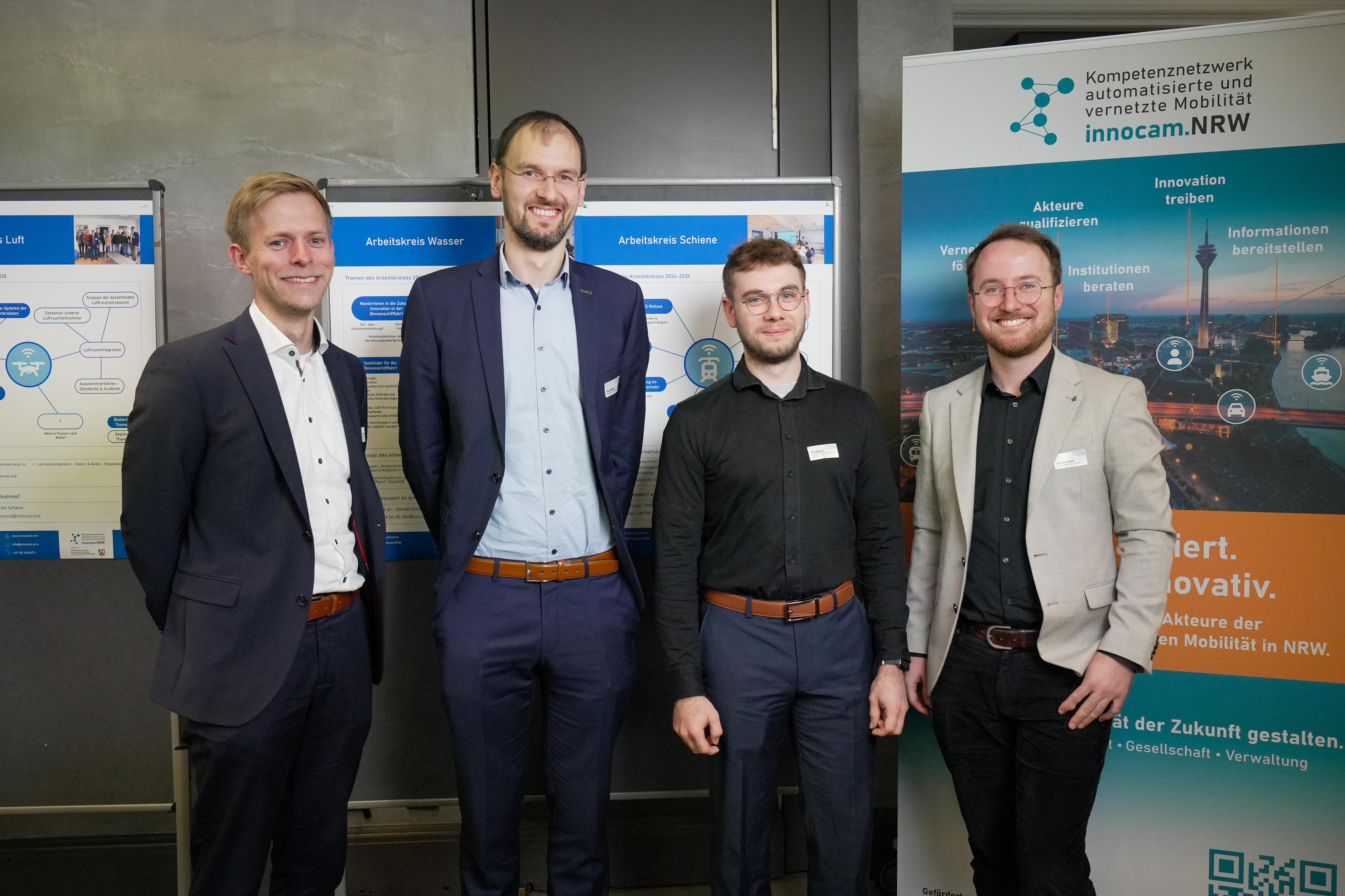 Vier Männer in Business-Kleidung stehen vor Informationsplakaten und einem Roll-up mit dem Logo und Text von innocam.NRW Mobility Meeting.
