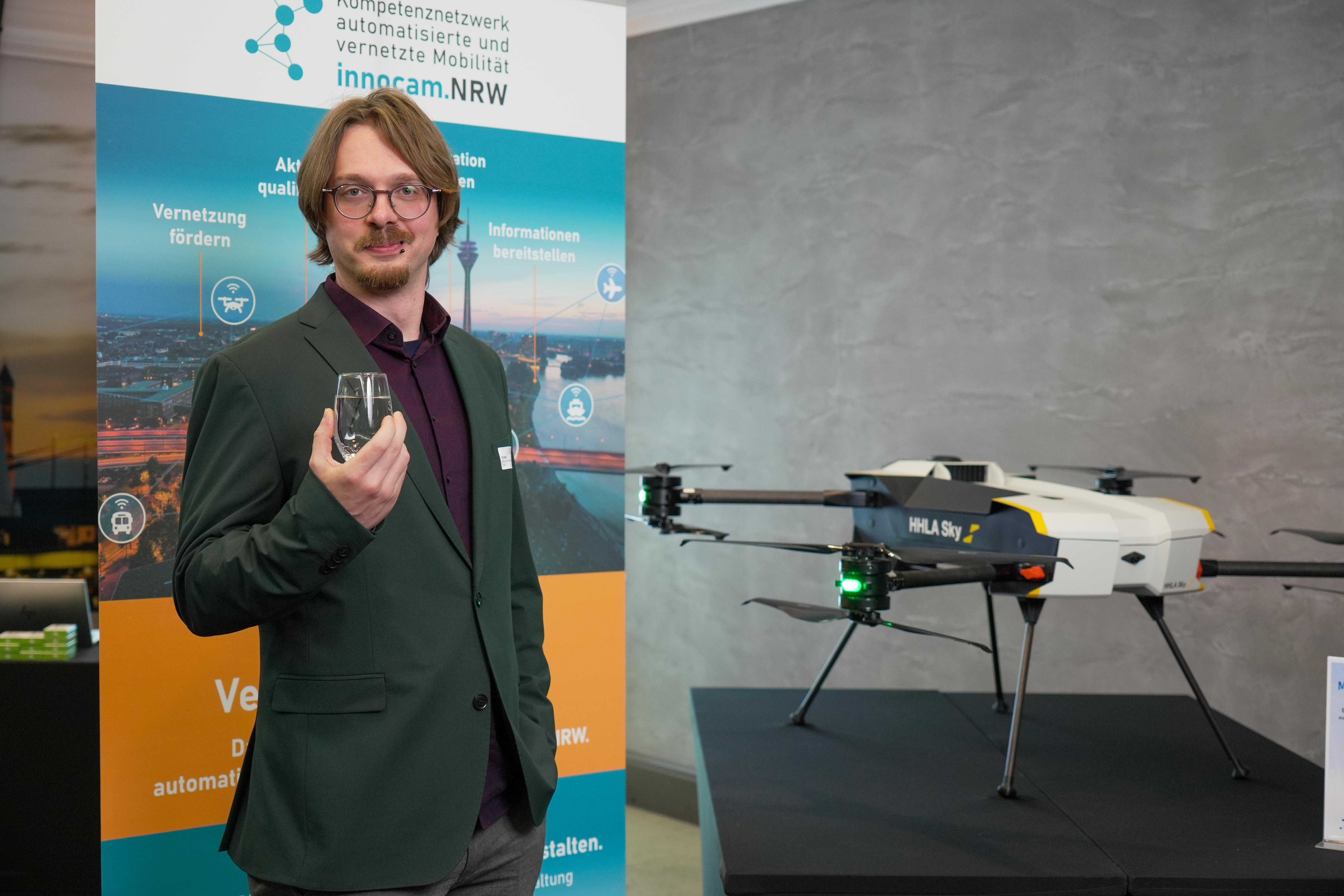 Mann in dunklem Sakko hält ein kleines Gerät, neben ihm steht eine Drohne mit der Aufschrift HHLA Sky auf einem schwarzen Podest, im Hintergrund ein Banner von innocam.NRW.