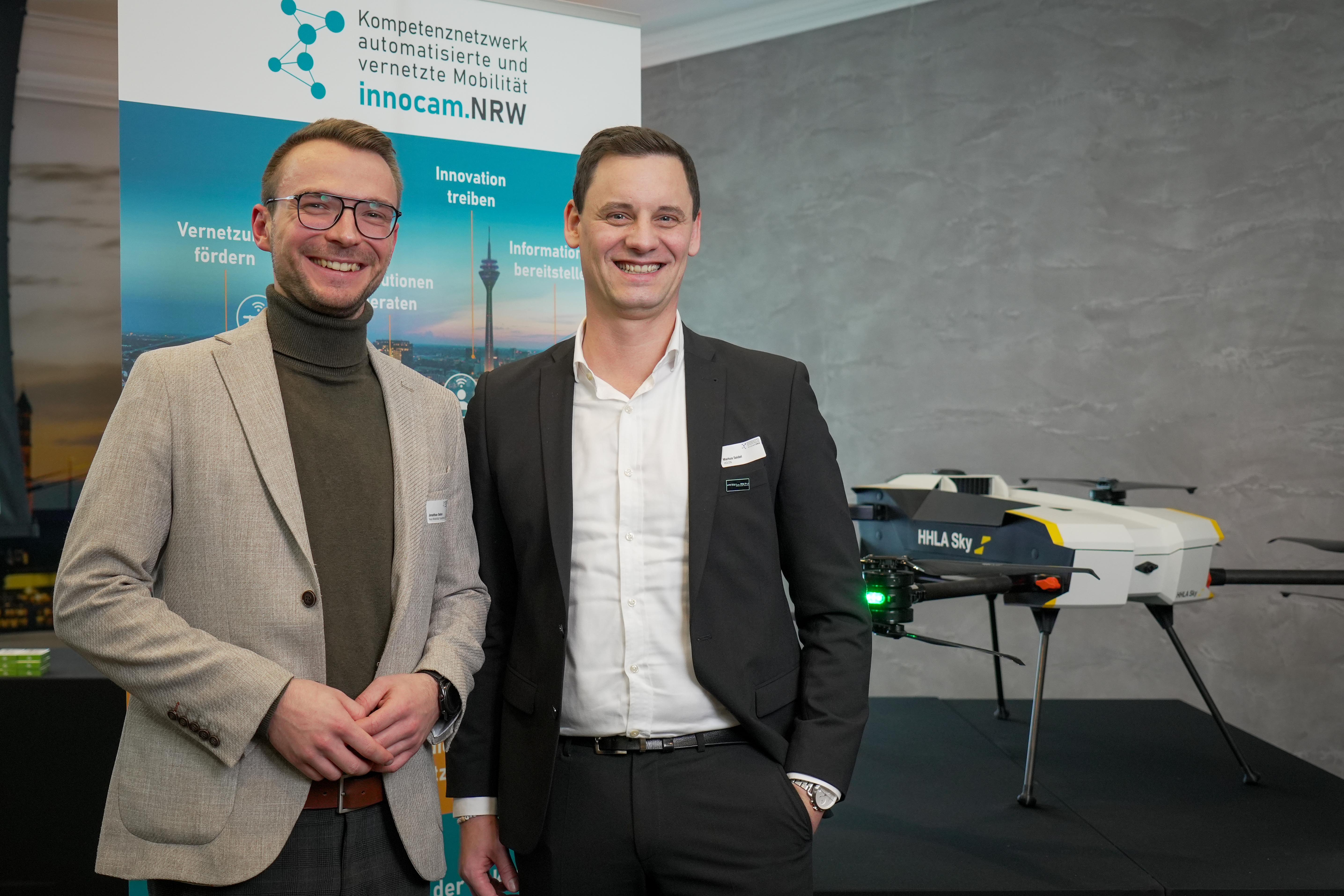 Zwei Männer in Businesskleidung stehen vor einem Banner mit dem Logo von innocam.NRW und einem weißen Drohnenmodell mit der Aufschrift 'HHLA Sky'.