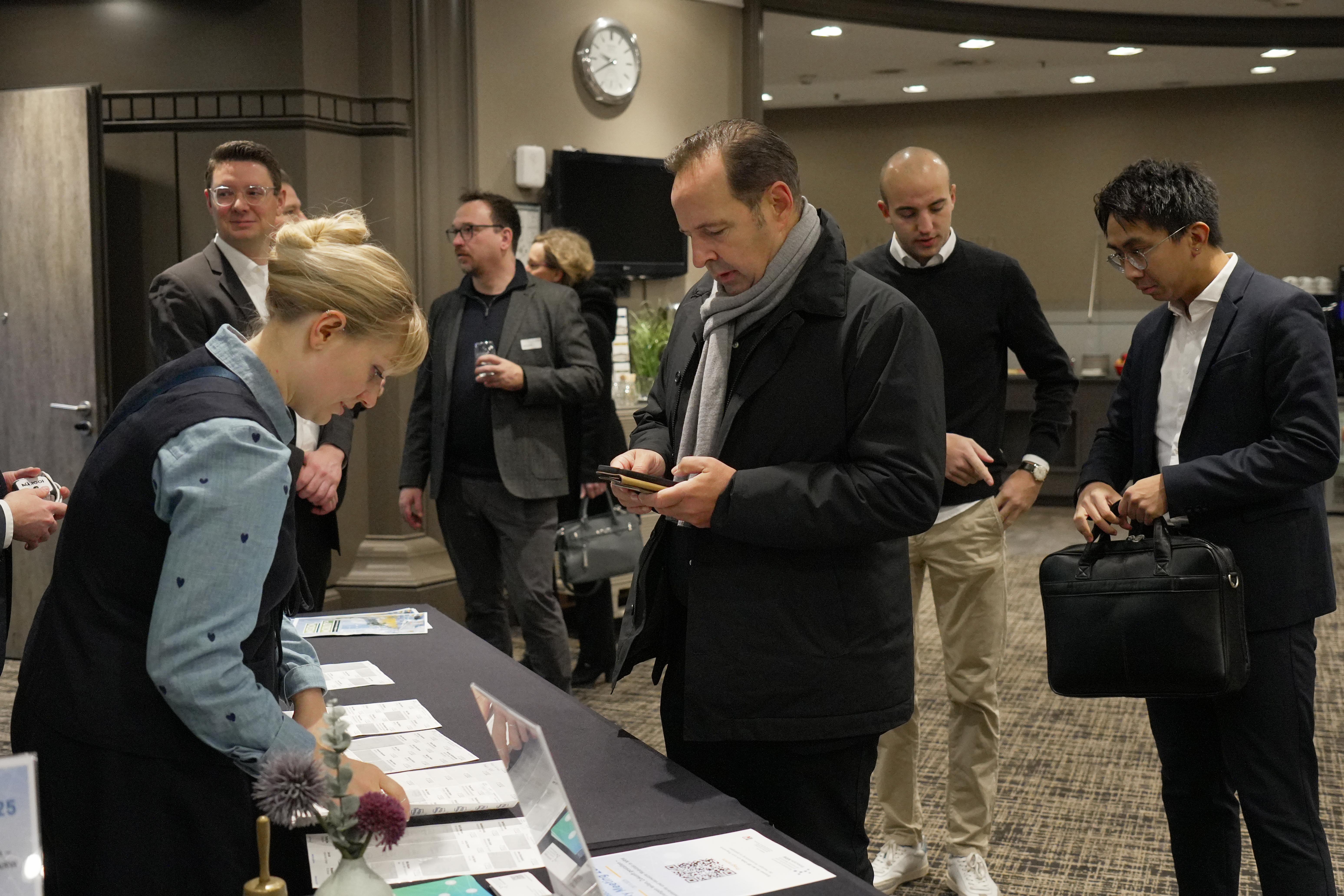 Mehrere Personen stehen an einem Registrierungstisch mit Informationsmaterialien bei einer Veranstaltung, einige nutzen Smartphones oder halten Taschen.
