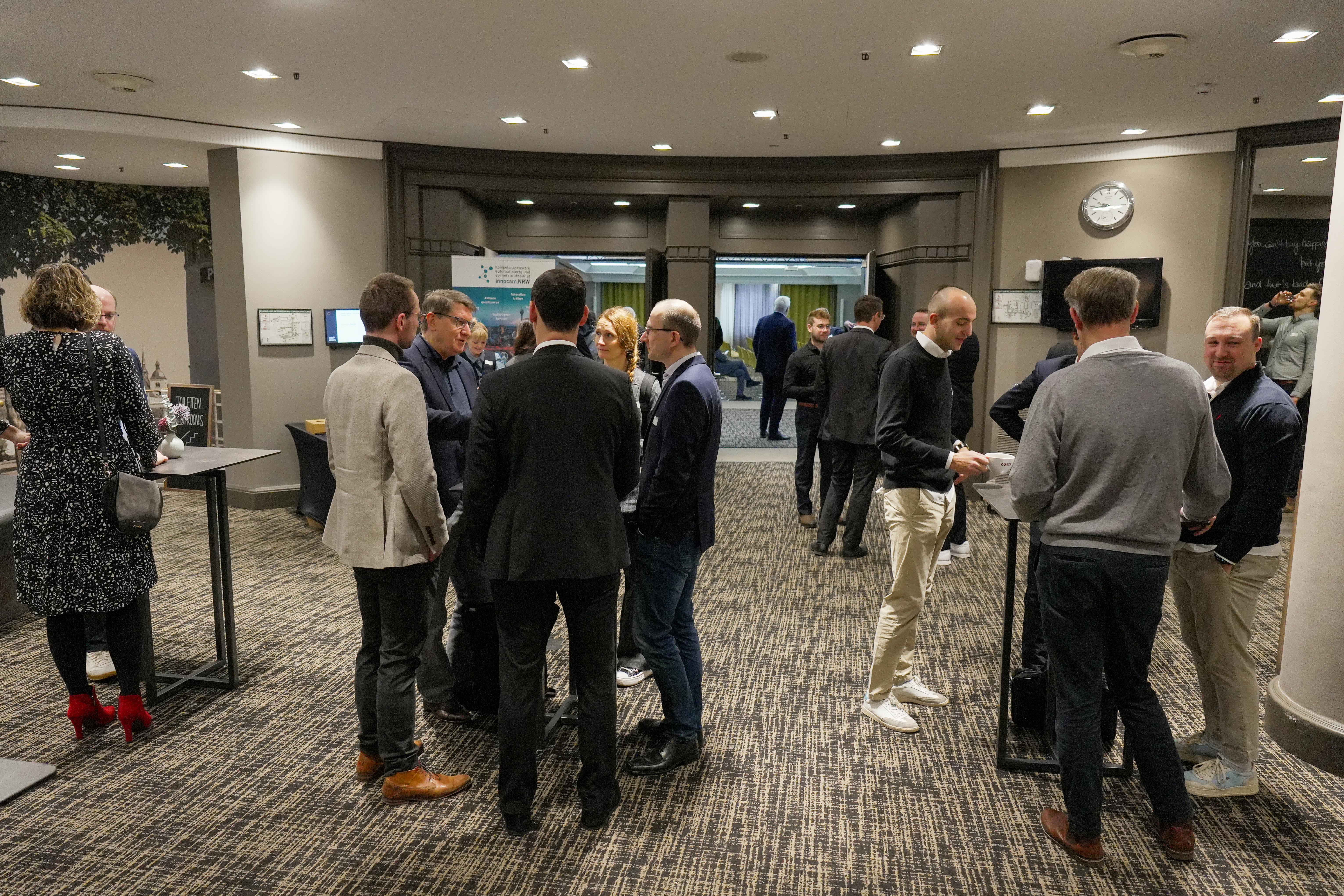 Mehrere Personen stehen in kleinen Gruppen in einem Konferenzraum an Stehtischen und unterhalten sich. 