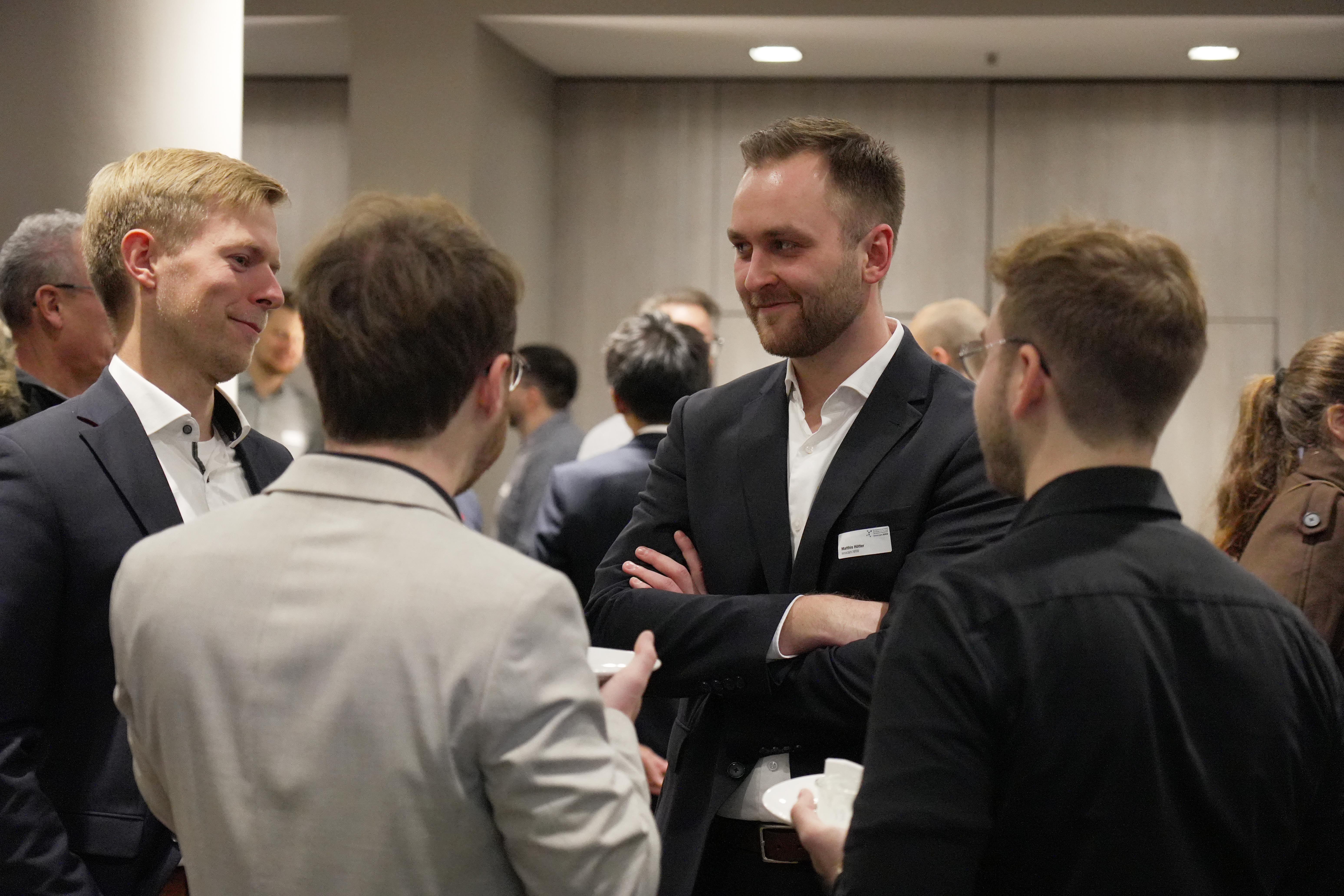 Mehrere Personen in Businesskleidung unterhalten sich in einem Innenraum bei einer Veranstaltung, im Vordergrund vier Männer.