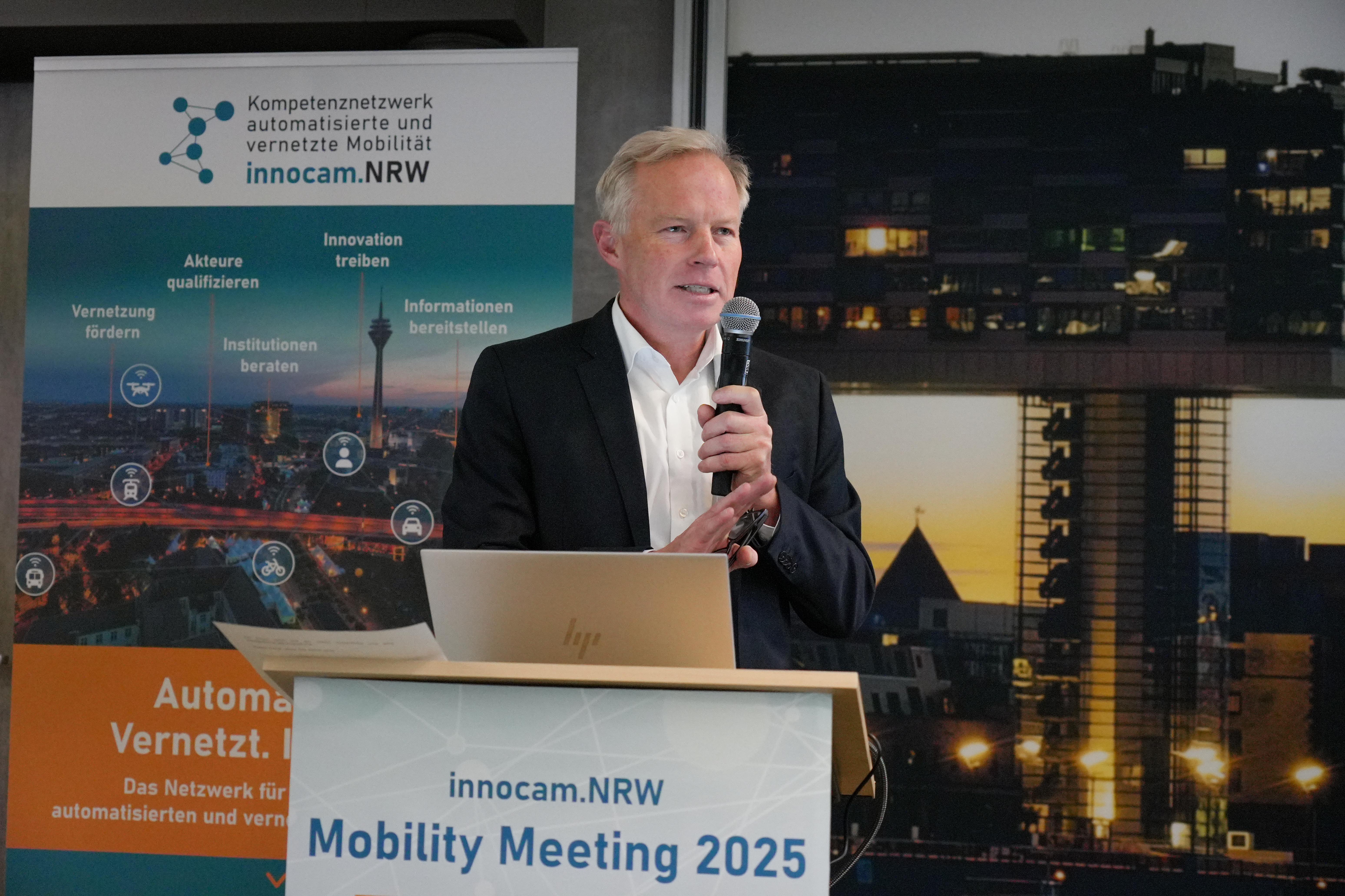 Mann in Anzug steht hinter einem Rednerpult mit Mikrofon, im Hintergrund Banner und Schild mit Aufschrift 'innocam.NRW Mobility Meeting 2025'.