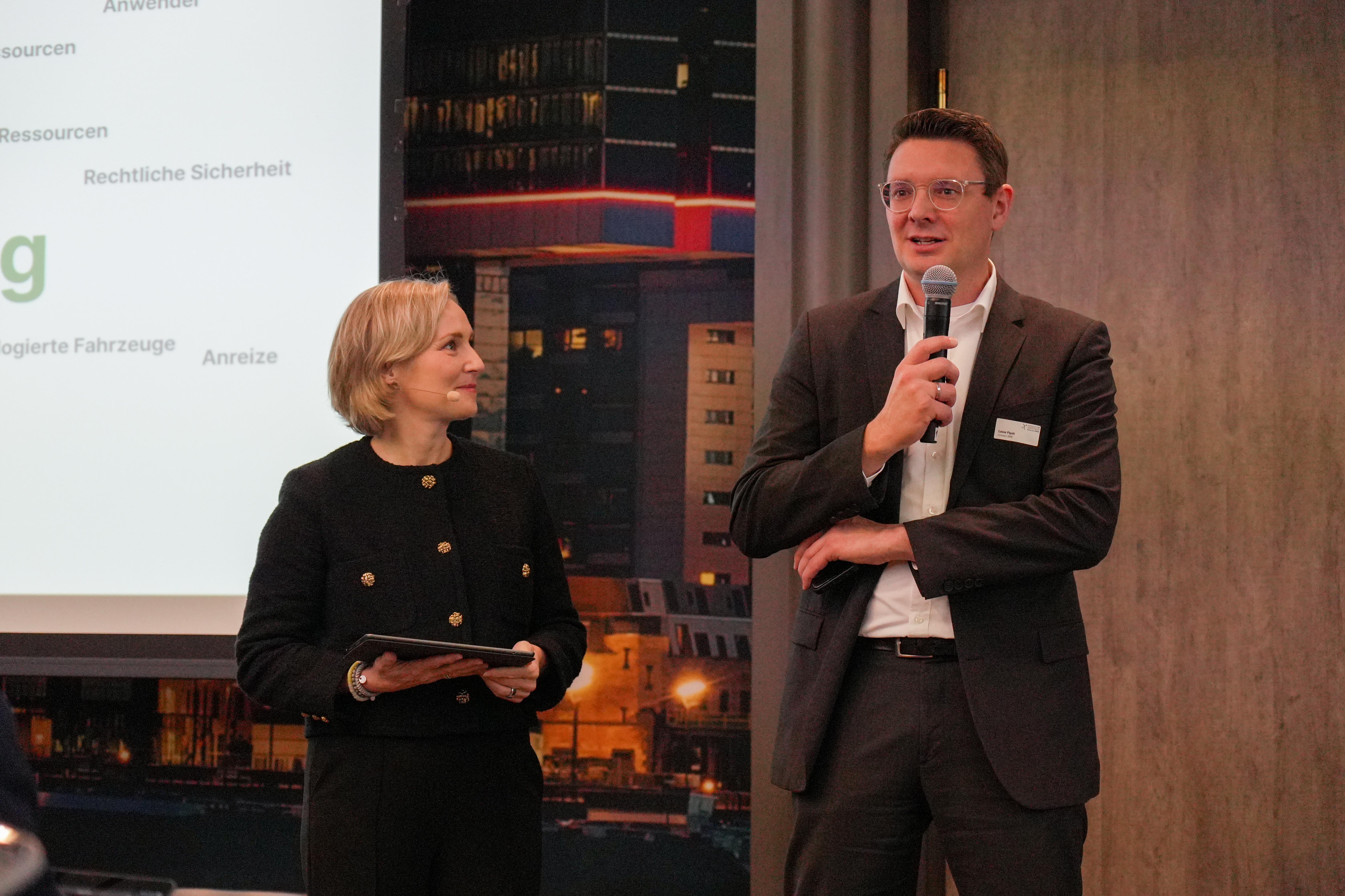 Zwei Personen stehen vor einer Präsentationswand, eine hält ein Mikrofon, die andere ein Tablet, beide tragen formelle Kleidung.