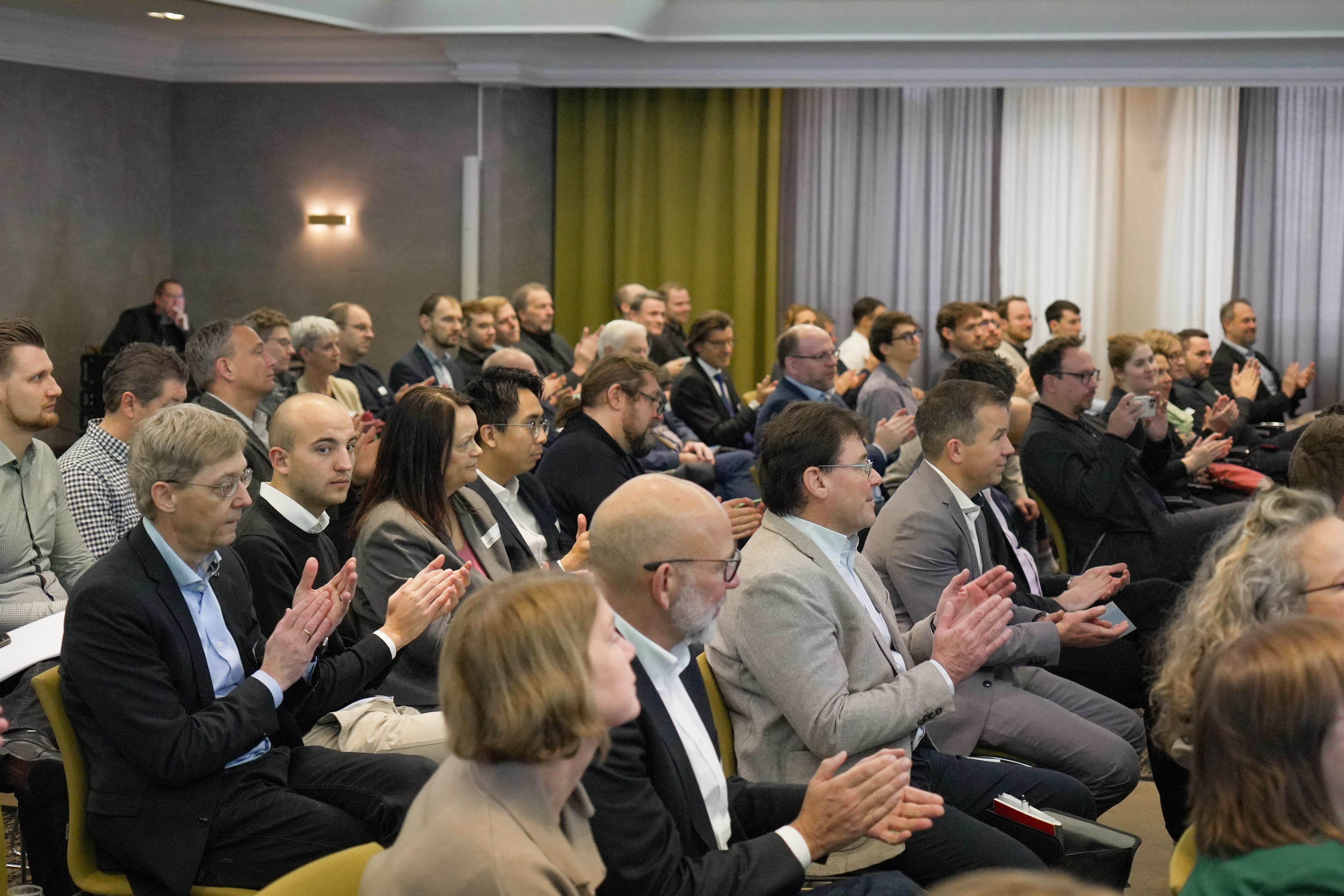 Mehrere Personen sitzen in Reihen in einem Konferenzraum und klatschen, einige tragen Anzug oder Business-Kleidung.