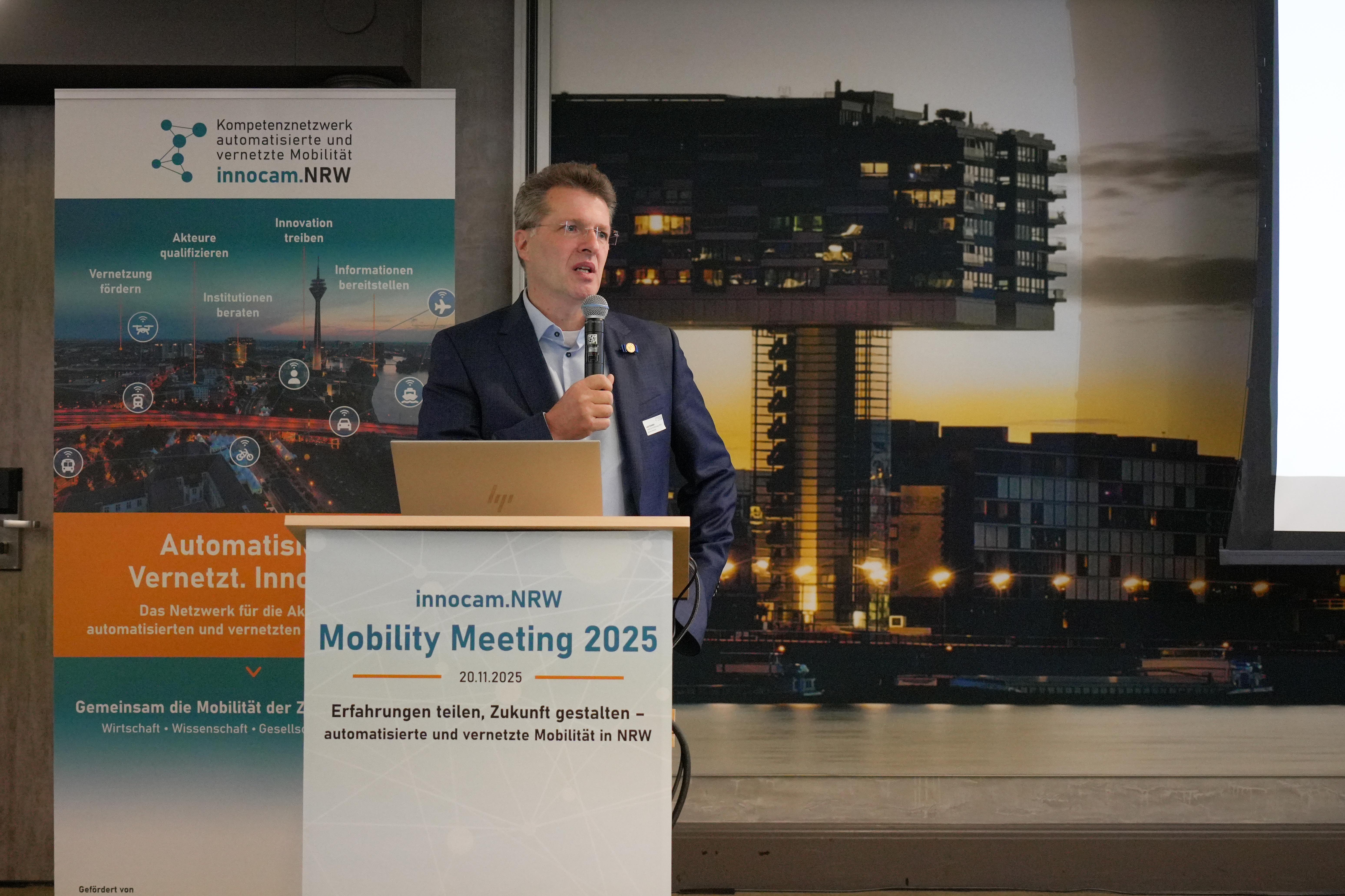 PRof. Dr. Eckstein hält Mikrofon und steht hinter Rednerpult mit Schild 'innocam.NRW Mobility Meeting 2025'. Im Hintergrund Banner mit Text und Grafiken zur automatisierten, vernetzten Mobilität.
