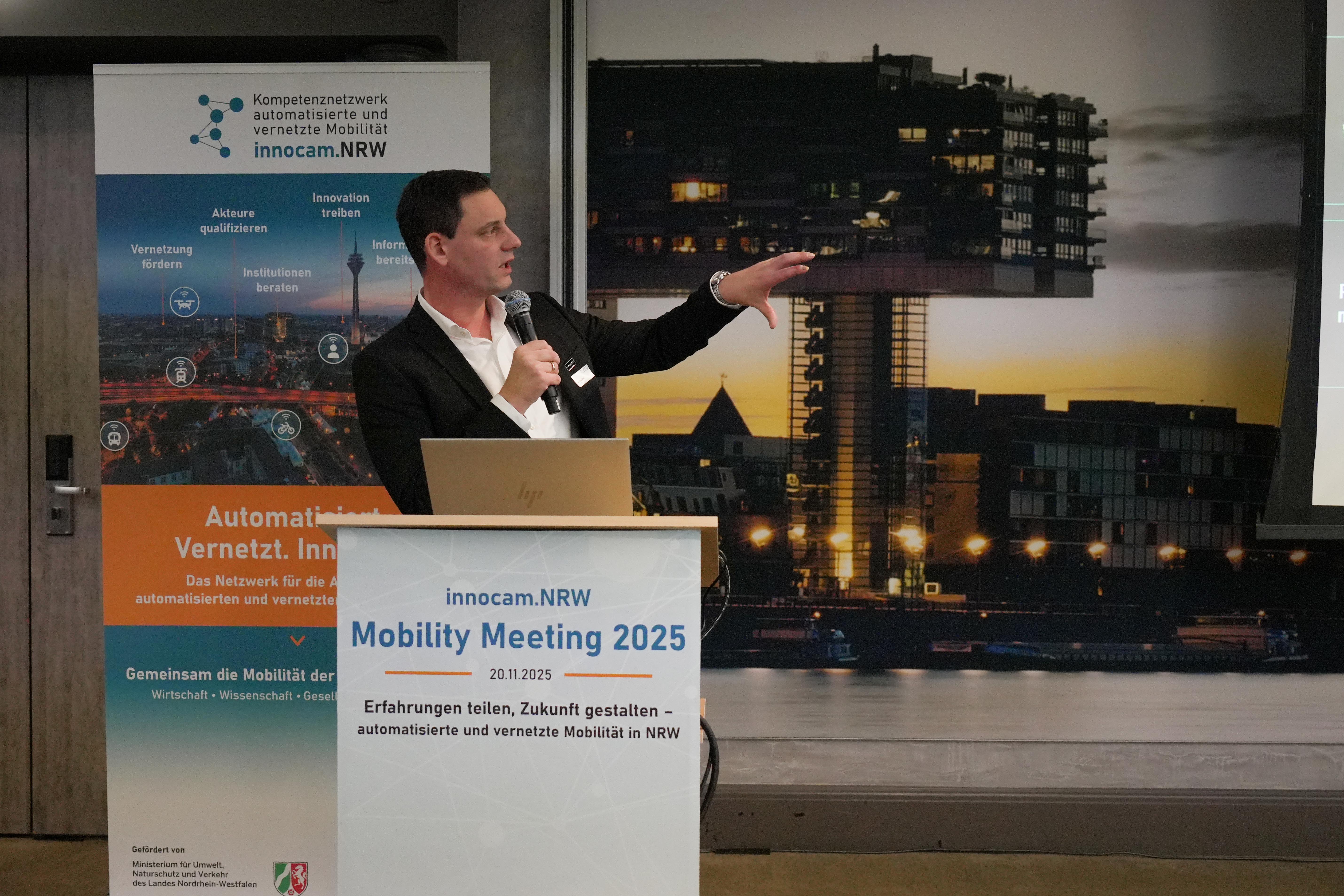 Mann mit Mikrofon und Sakko steht am Rednerpult mit Schild 'innocam.NRW Mobility Meeting 2025' vor Plakat und großem Bild eines modernen Gebäudes bei Nacht.