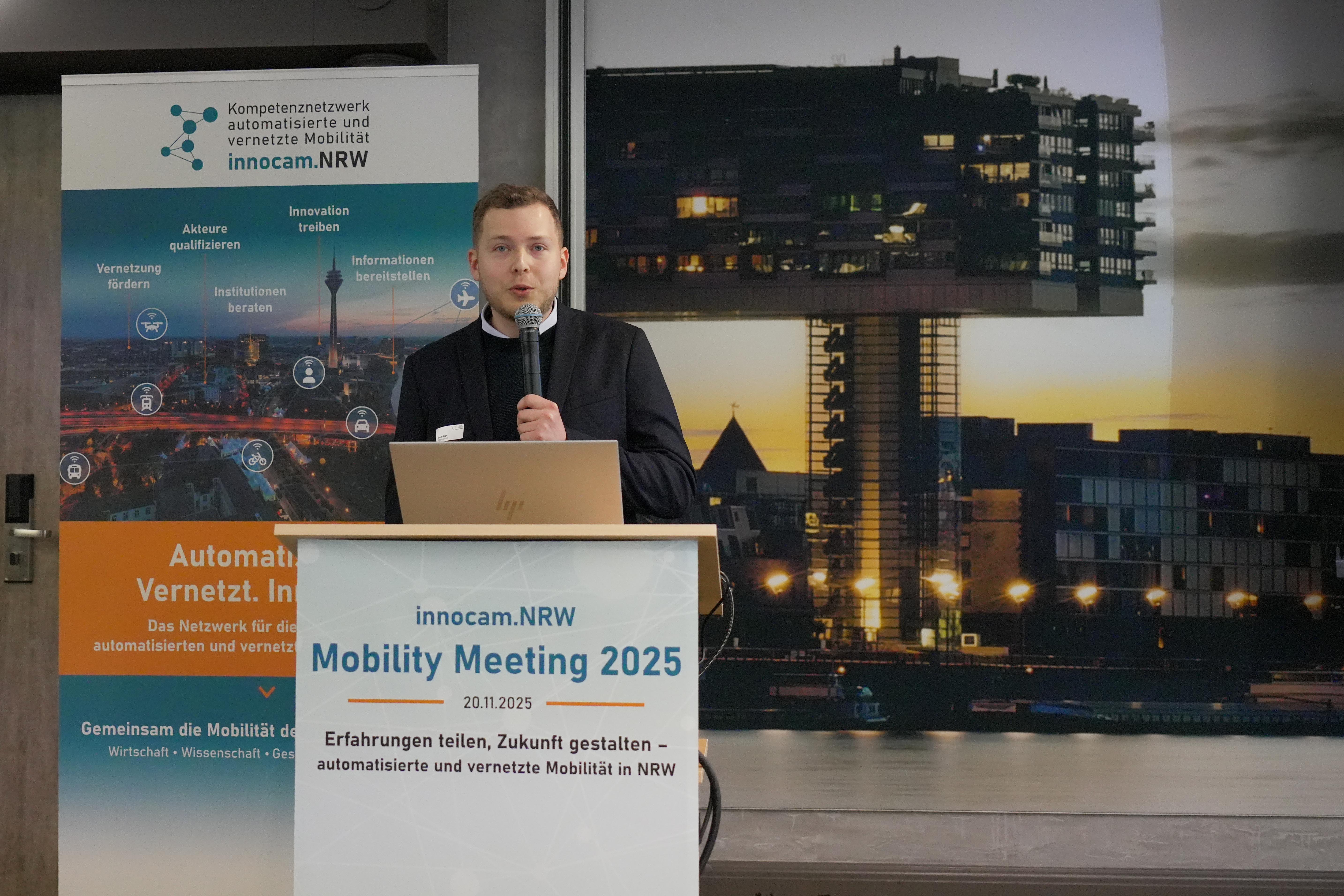 Person steht an einem Rednerpult mit Mikrofon und Laptop, im Hintergrund Banner und großes Bild eines modernen Gebäudes bei Dämmerung.