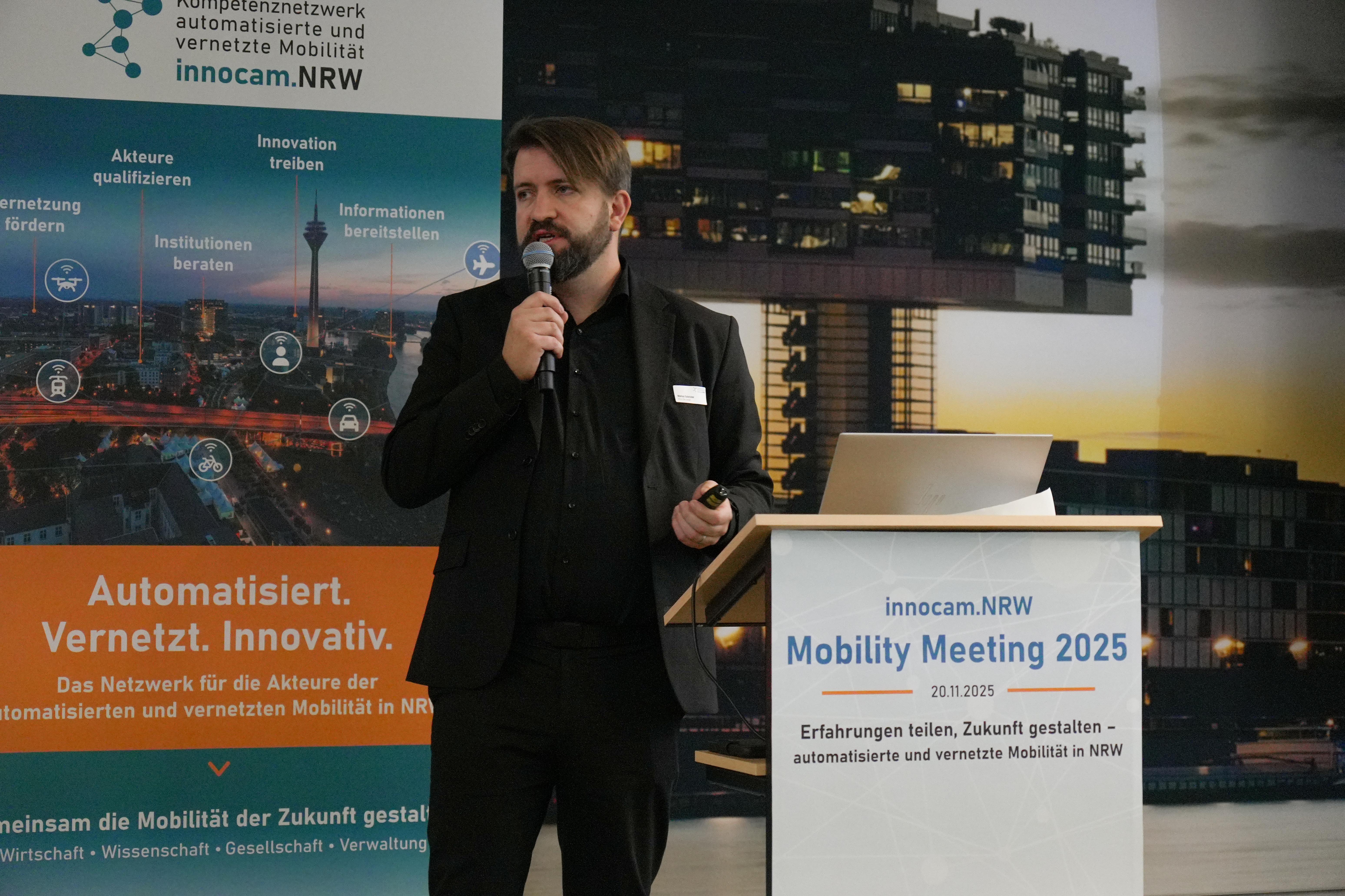 Ein Mann in dunkler Kleidung hält ein Mikrofon und steht vor einem Rednerpult mit einem Schild 'innocam.NRW Mobility Meeting 2025'. Im Hintergrund sind ein Banner mit Text und ein großes Bild eines Gebäudes zu sehen.