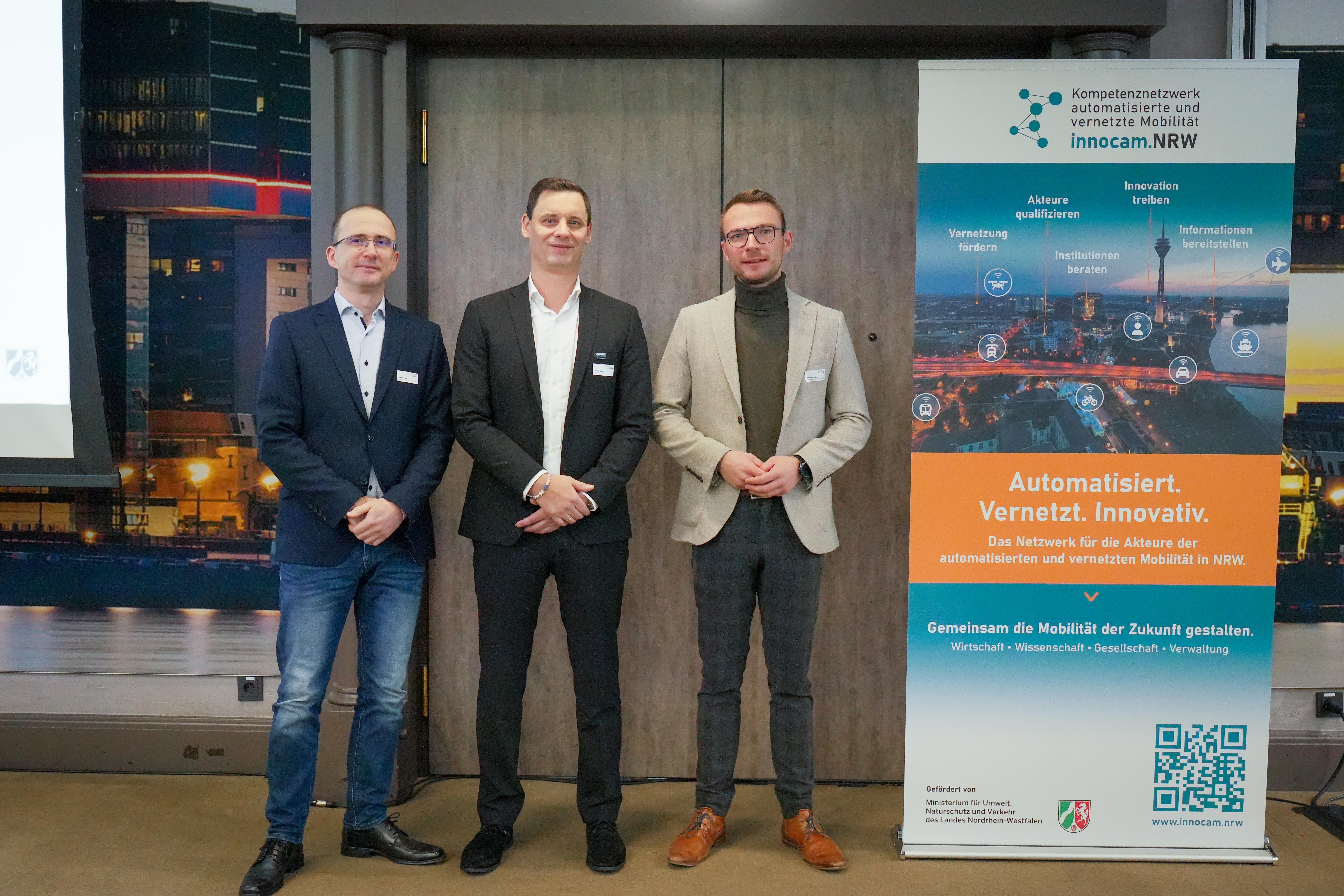 Drei Männer in Business-Kleidung stehen vor einer Tür neben einem Roll-up mit dem Titel 'innocam.NRW' und dem Slogan 'Automatisiert. Vernetzt. Innovativ.'