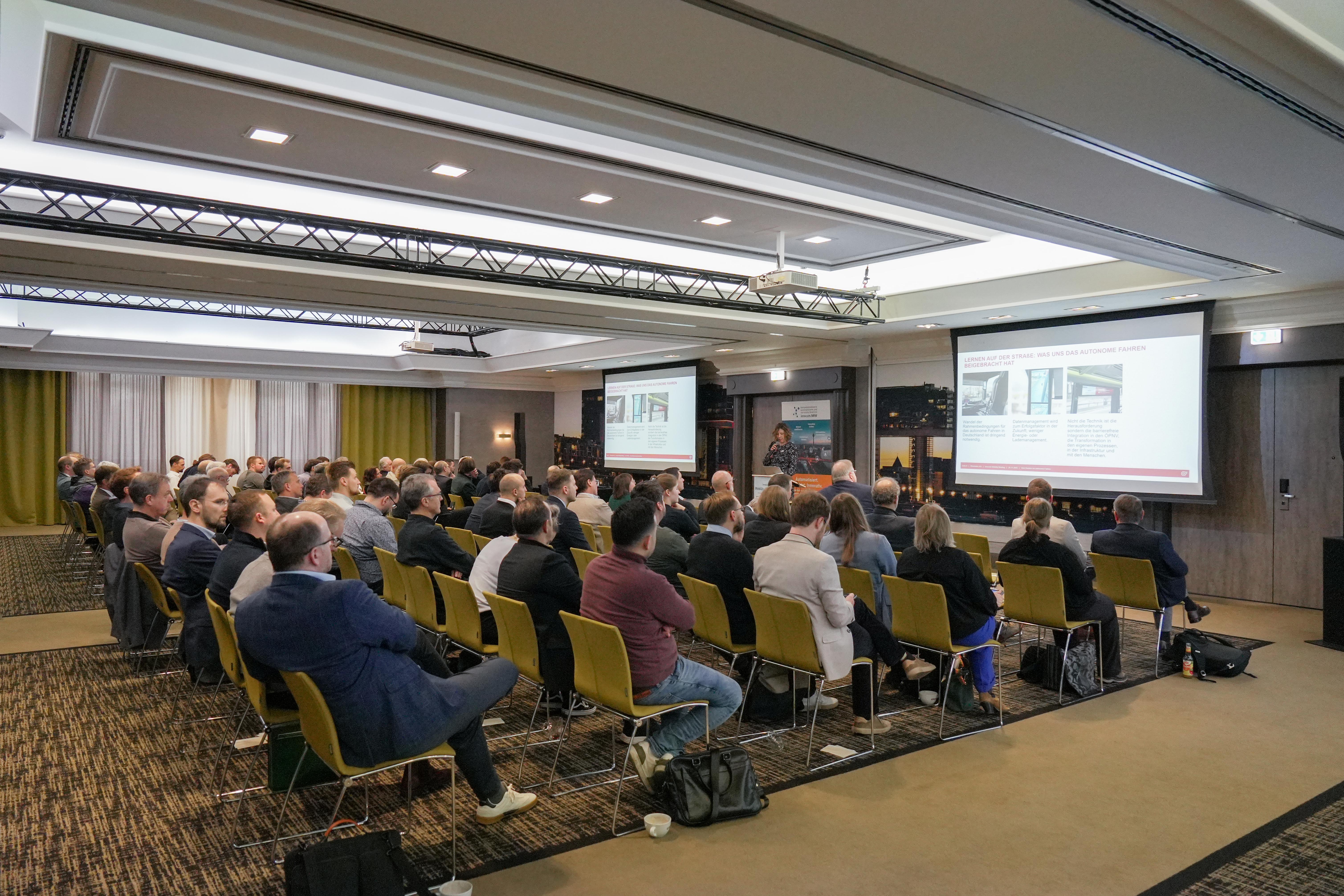Konferenzraum mit mehreren Personen, die auf zwei Projektionsflächen eine Präsentation verfolgen, einige tragen Business-Kleidung, Sitzreihen mit gelben Stühlen.