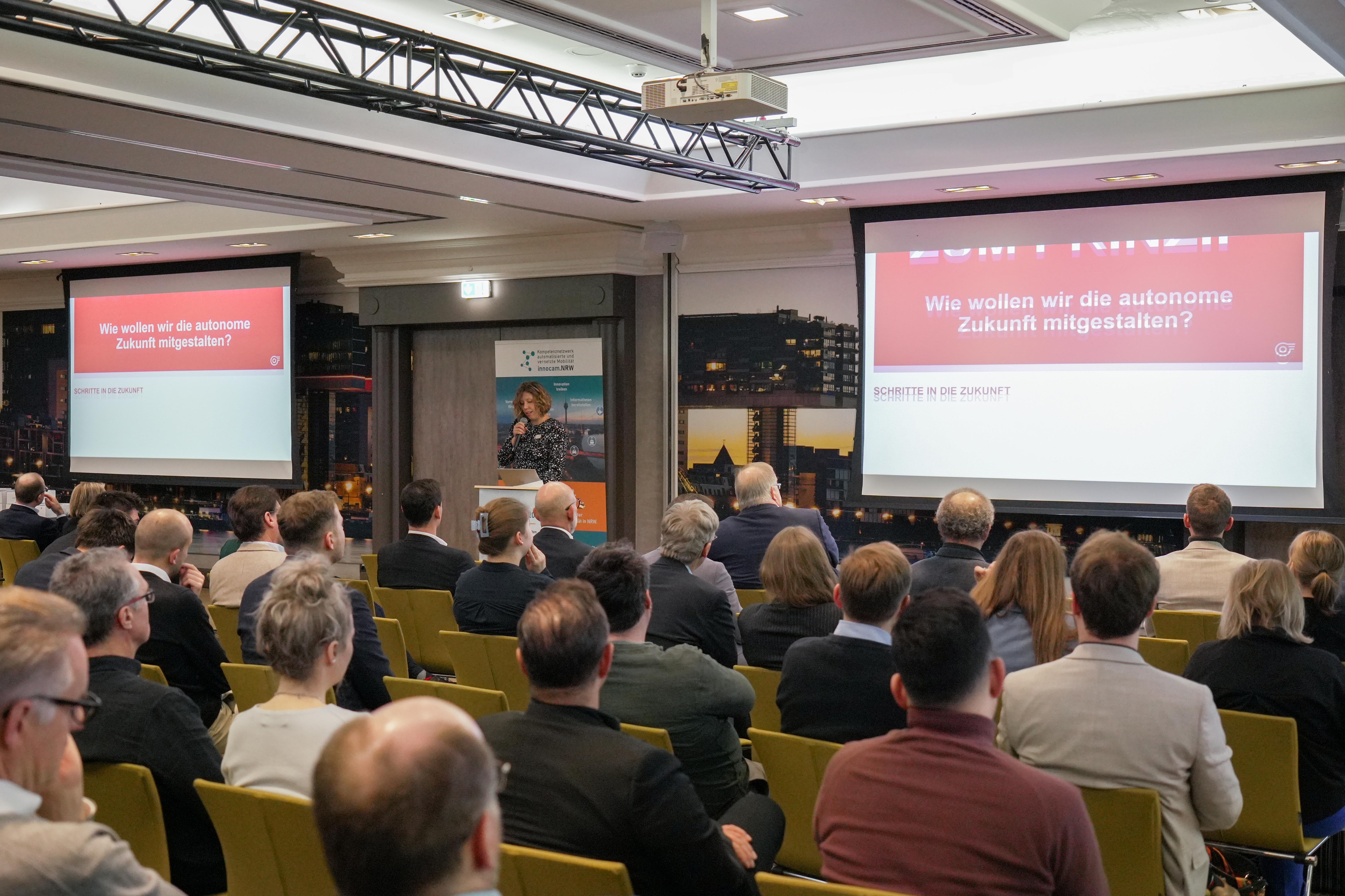 Konferenzraum mit Publikum, das einer Präsentation zum Thema autonome Zukunft folgt, sichtbar auf zwei Projektionsflächen mit dem Text 'Wie wollen wir die autonome Zukunft mitgestalten?'.