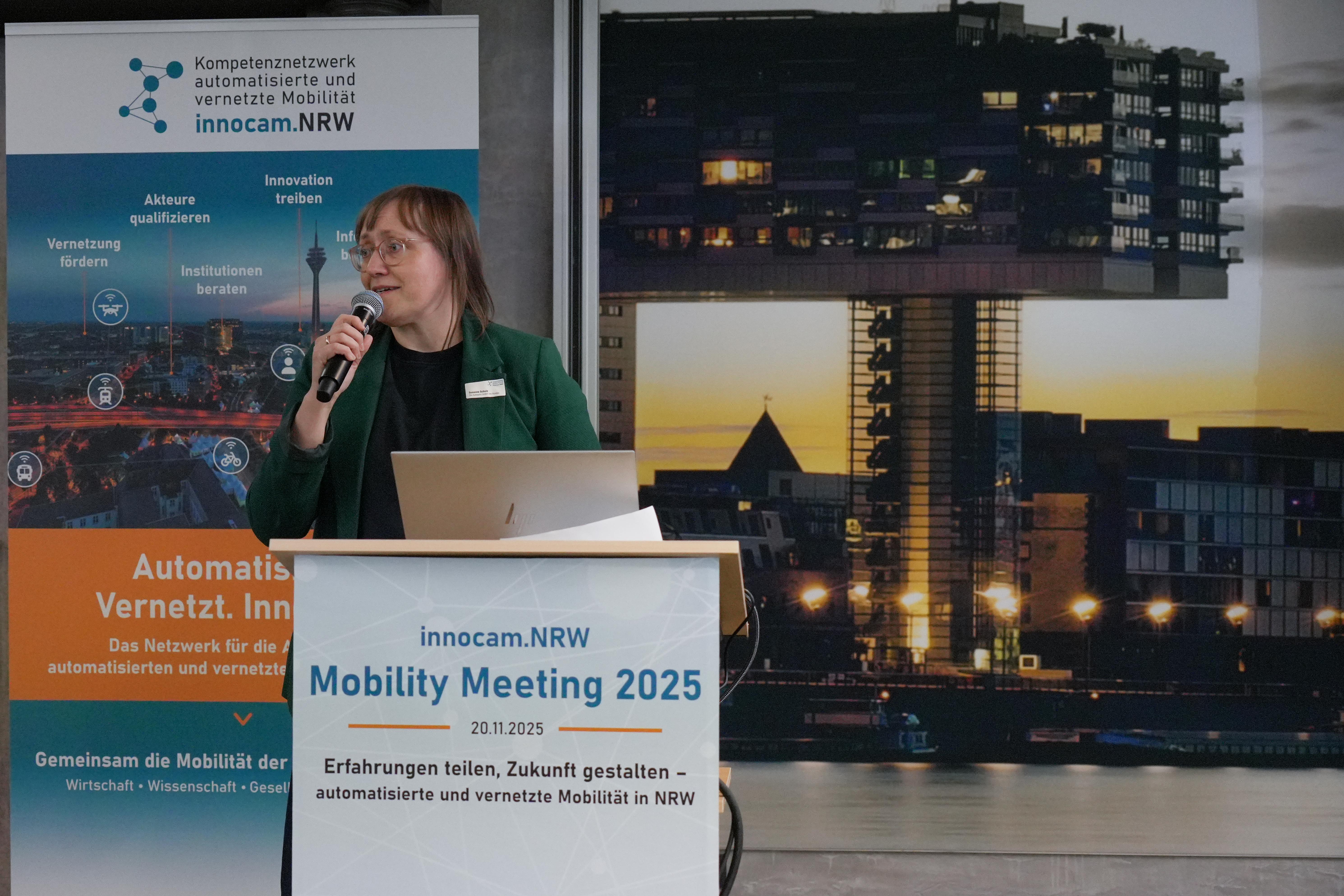 Frau mit Mikrofon steht an einem Rednerpult mit Schild 'innocam.NRW Mobility Meeting 2025' vor einem Banner des Kompetenznetzwerks automatisierte und vernetzte Mobilität und einem nächtlichen Stadtpanorama mit beleuchteten Gebäuden.