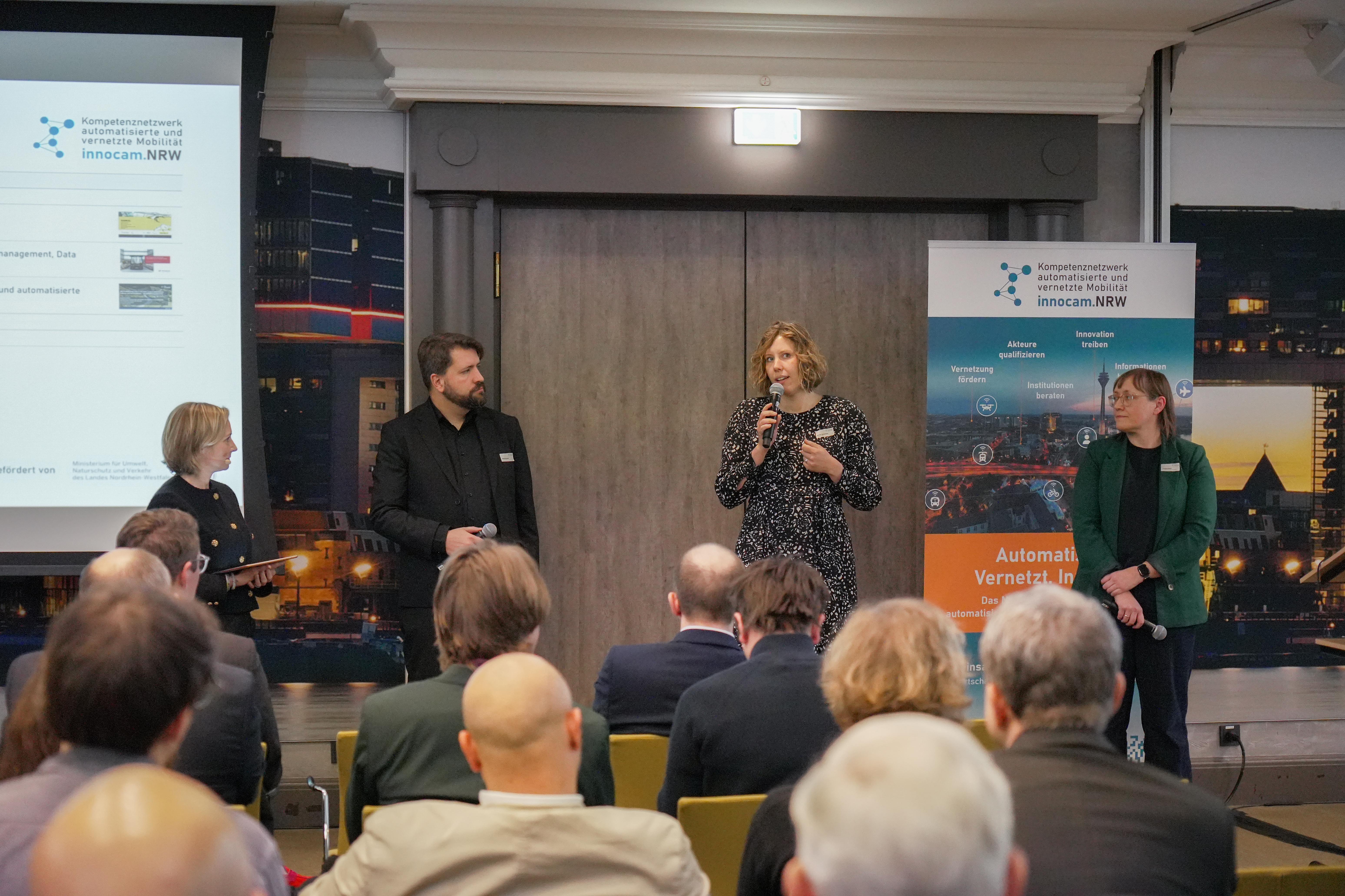 Vier Personen stehen vor einem Publikum in einem Konferenzraum, zwei Banner mit dem Logo und Text von innocam.NRW sind im Hintergrund sichtbar.