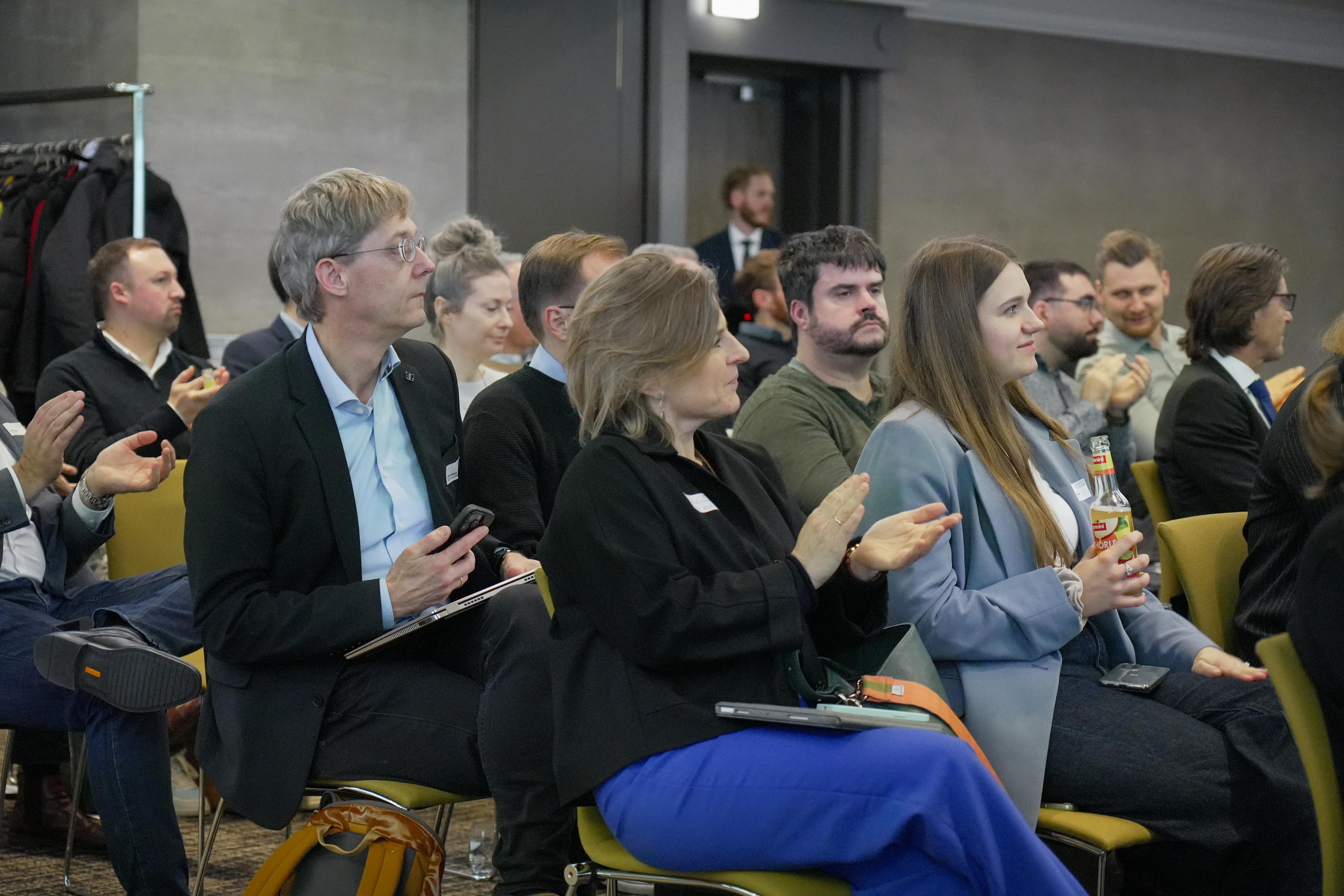 Mehrere Personen sitzen in einem Raum bei der innocam.NRW Mobility Meeting Veranstaltung, einige klatschen, eine Frau hält eine Flasche, andere haben Notizblöcke oder Smartphones in der Hand.