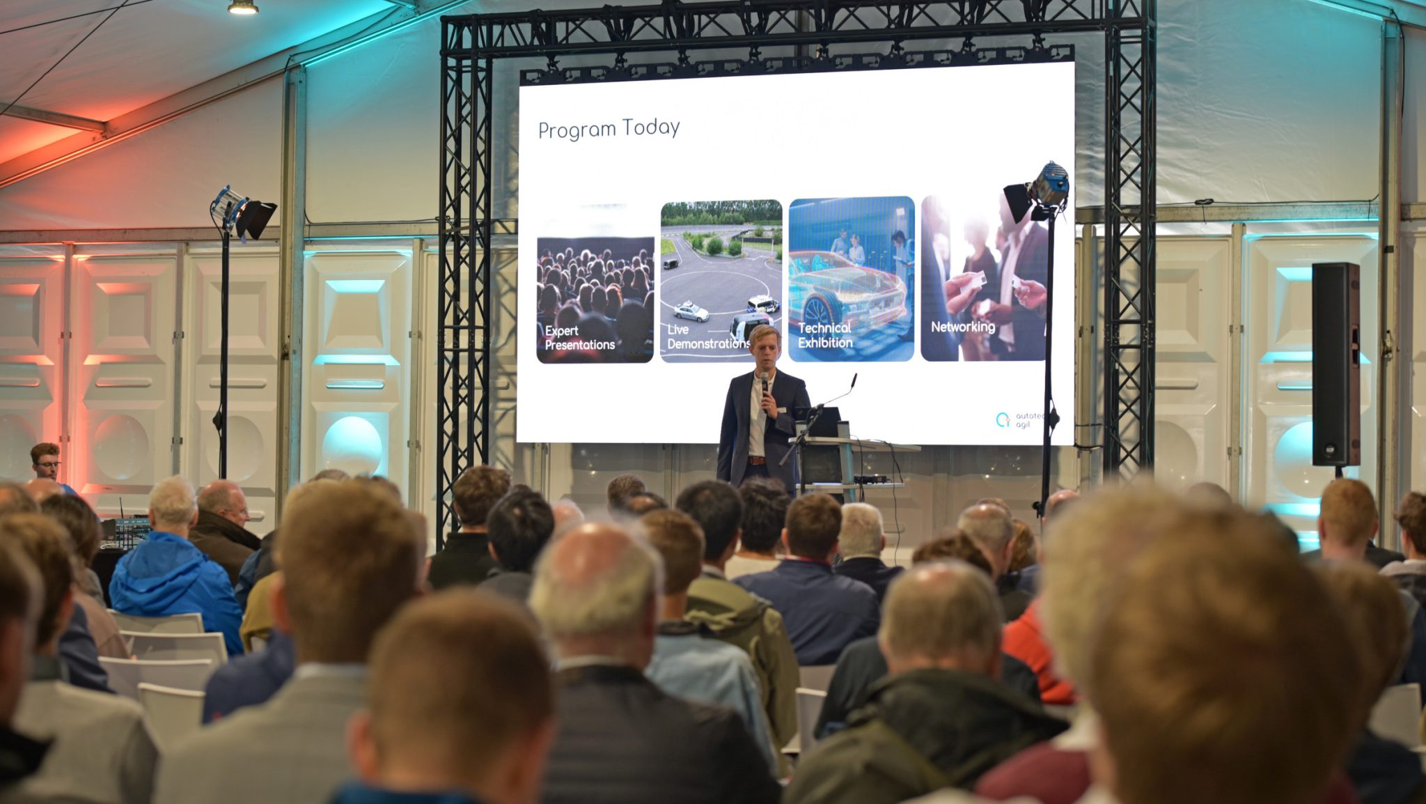 „Eine Person hält eine Präsentation vor einem Publikum in einem Konferenzzelt. Auf der großen Leinwand hinter ihr steht ‚Program Today‘ mit vier Themen: Expert Presentations, Live Demonstrations, Technical Exhibition und Networking. Die Veranstaltung hat ein professionelles Umfeld mit Beleuchtung und Bühnenaufbau.
