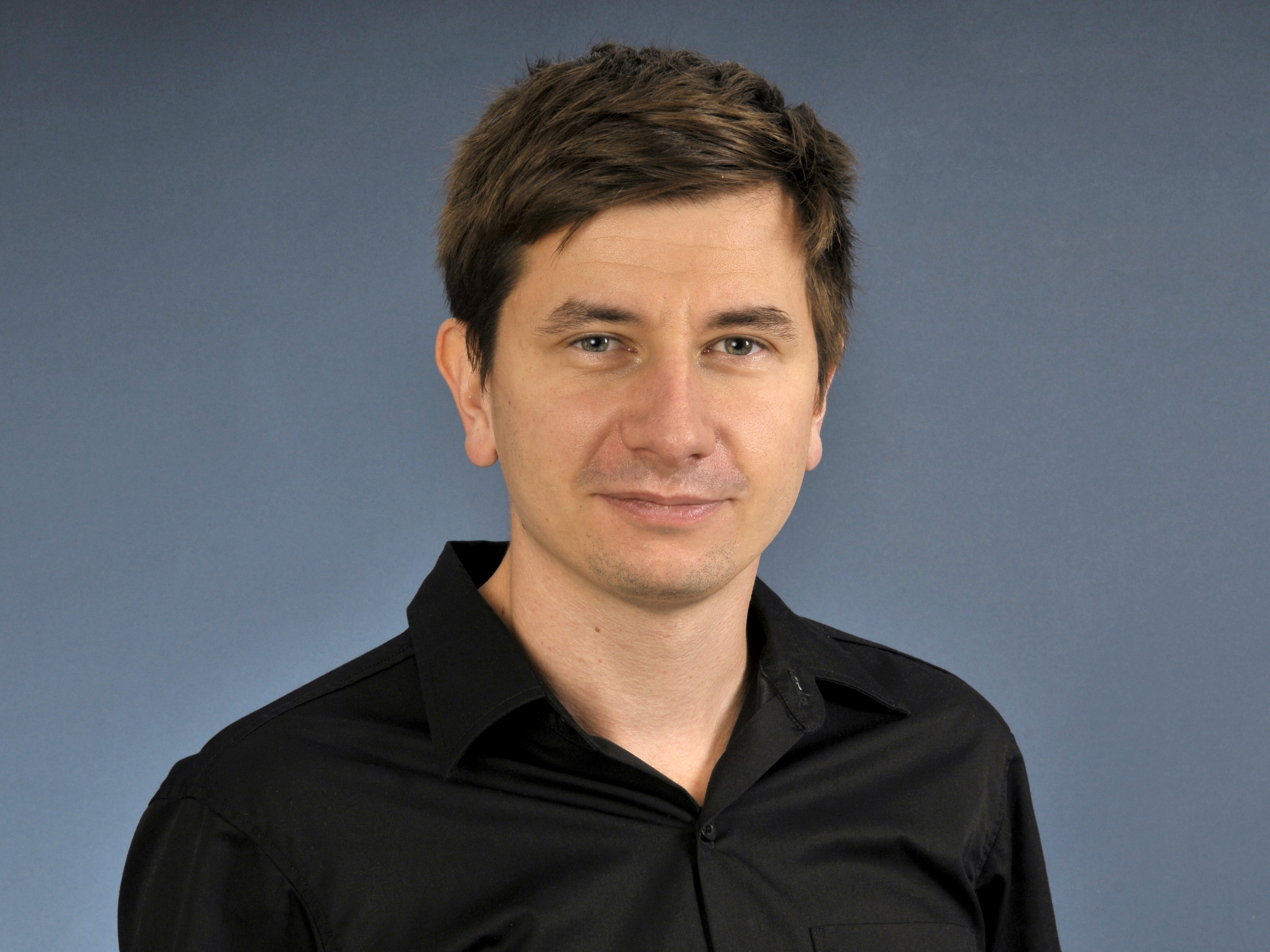 Portrait von Manuel Cymbaly zeigt in in einem dunklen Hemd, lächelnd mit dunklem Haar vor einem dunkelblauen Hintergrund.